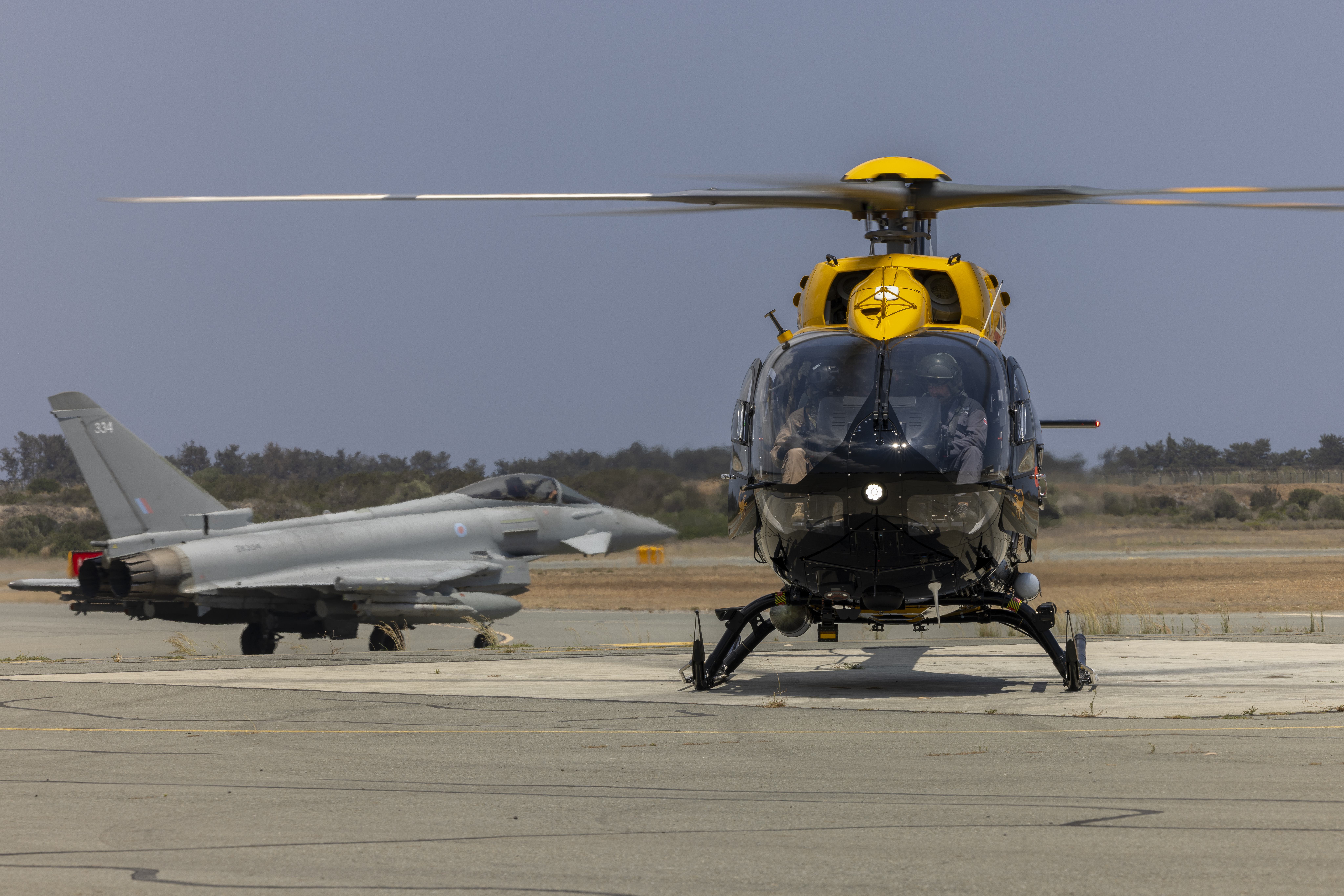 An H145 Jupiter helicopter has rotors running on the pan at RAF Akrotiri with a Typhoon taxiing in the background.