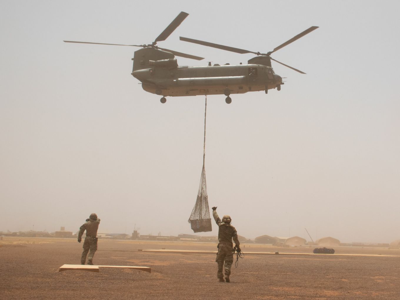 Chinook carries underslung load above personnel on ground.