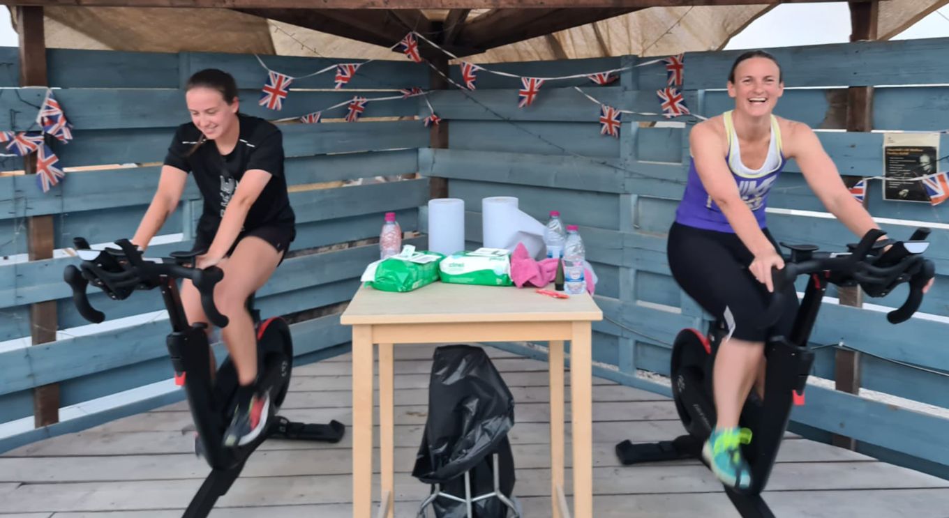 Two personnel on exercise bikes with hand sanitiser station between them.