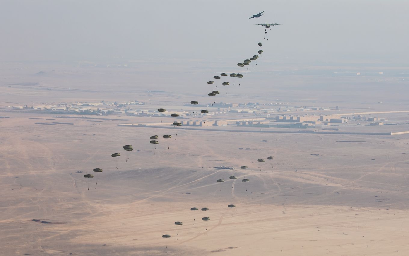 Two C-130J Hercules aircraft airdrop lots of parachuting troops over the desert.