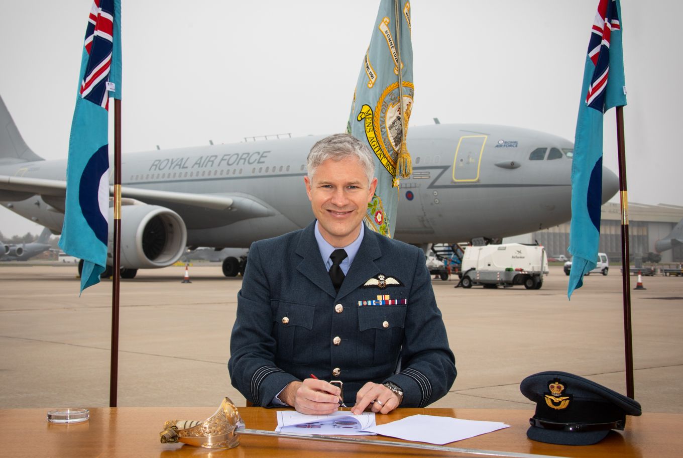 Wing Commander Udall signing a document at a table outside, with the Airbus Tanker Aircraft in the background. 