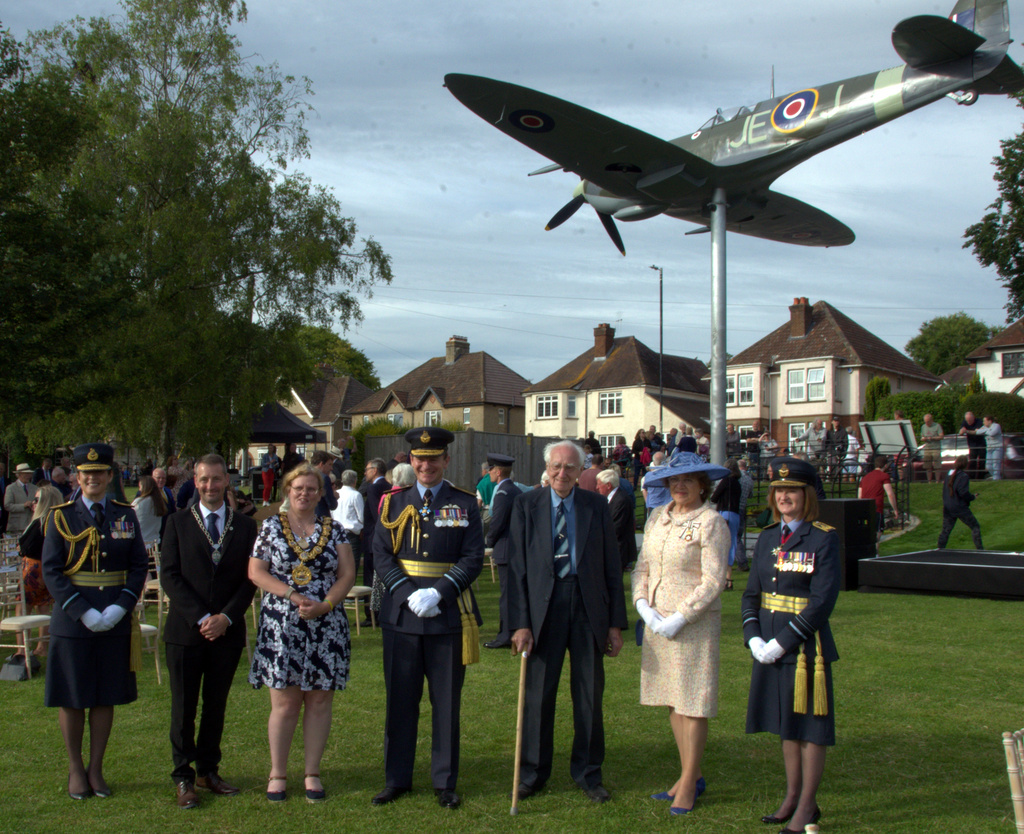 Seven notable people stand for a portrait with Spitfire model statue and members of the public.