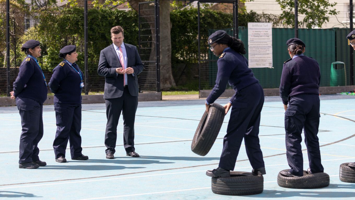 Female Cadet work together moving tires, while teachers and James Heappey watch on.