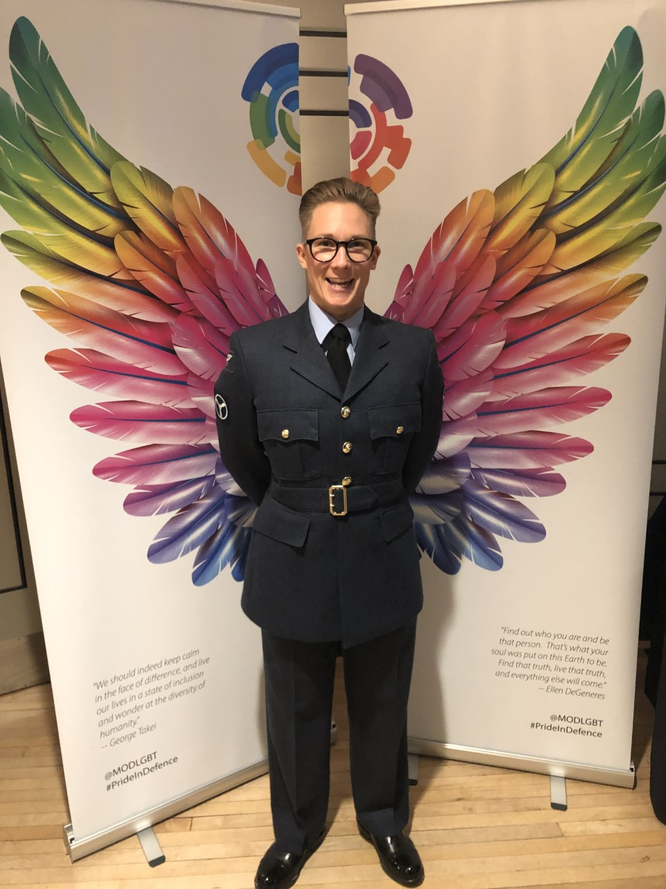 Senior Aircraftman Em Holness stands in-front of rainbow coloured wings artwork.
