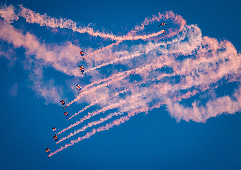 Decoration: Photo - Having jumped from the RAF Globemaster (C-17A) display aircraft, beneath red, white and blue rectangular parachutes. Each parachute is emblazoned with the huge black letters, 'R A F', one letter on each of the white stripes. Descending in a loose 'stack' of 9 team members they have moved from right to left, streaming pink smoke from cannisters on their legs.