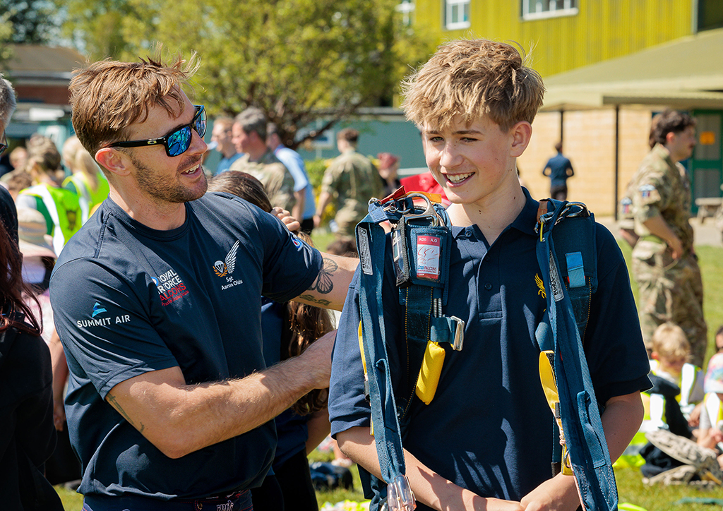 Decoration: Photo - In the foreground, experienced second year Falcon, Sergeant Aaron Olds fits a parachute harness to an onlooker, who wishes to feel the experience of wearing it. In the immediate background is a crowd of people and, beyond them is the Station Gym.