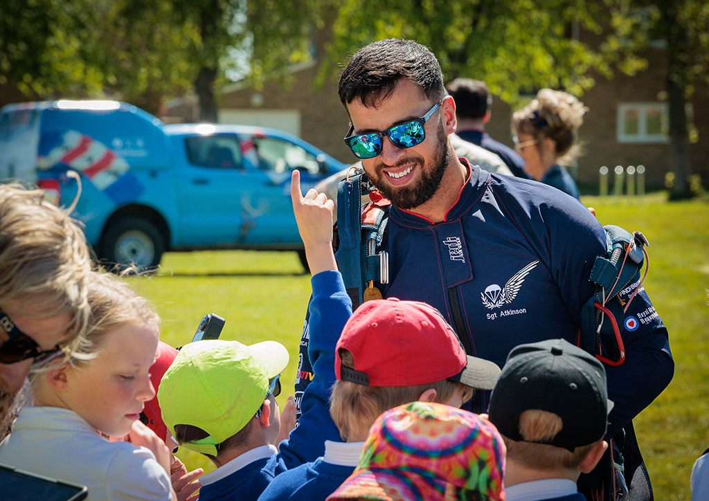 Decoration: Photo - In the foreground, experienced third year Falcon, Sergeant Atkinson, is seen here chatting with a group of school children who are asking him questions. Two adults are seen behind him on the right, and on the left is the RAF Falcons display vehicle, emblazoned with images of the Falcons and their parachutes.