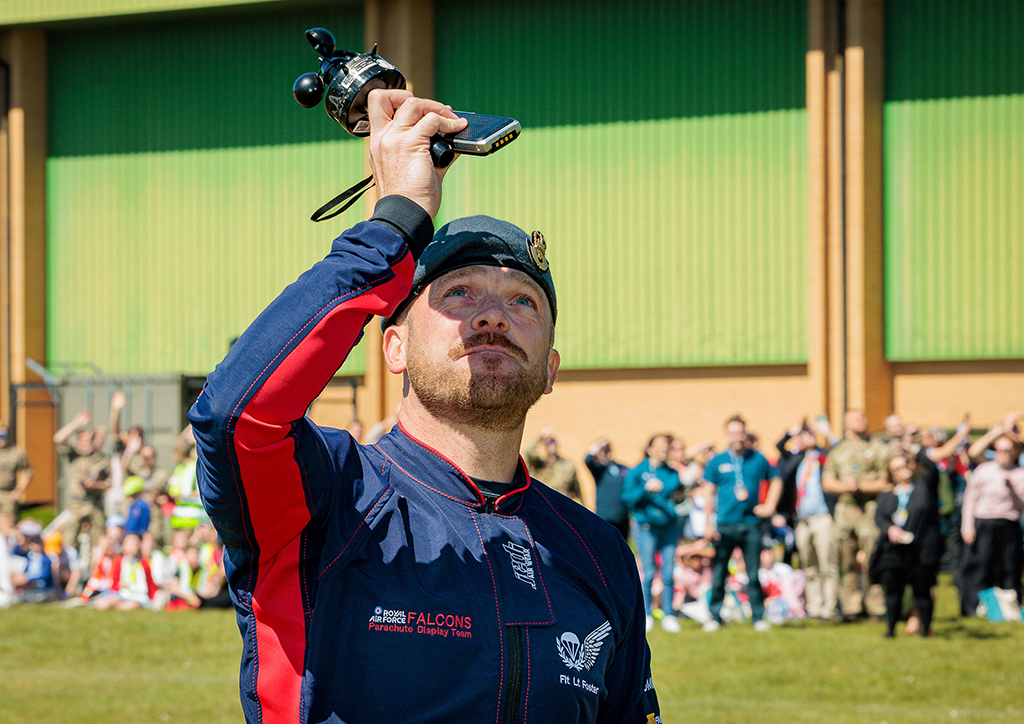 Decoration: Photo of Flight Lieutenant Adam Foster, Deputy Officer Commanding RAF Falcons, stands in the sports field drop zone, with the Station Gym and crowds of people in the background, while using instruments to ensure that the weather conditions are within limits and it is safe for the Falcons to begin their display.