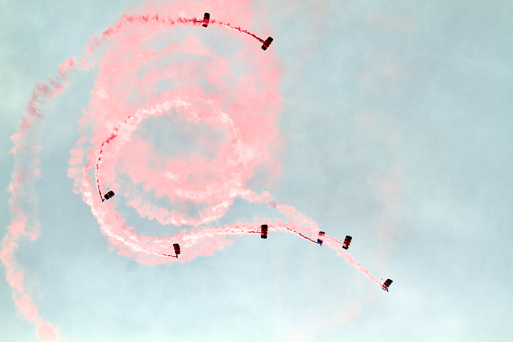 An aerial view of RAF Falcons Parachute Display Team performing manoeuvres in a cloudy sky, with vibrant pink smoke trails that form large spiral patterns behind the eight parachutists.