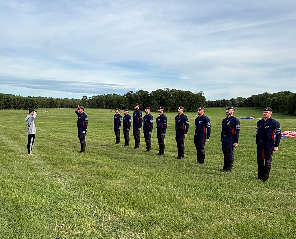 A row of nine RAF Falcons Parachute Display Team in dark blue and black uniforms wearing berets stand in formation behind their coach on a grassy field, saluting a person standing opposite them under a partly cloudy sky.