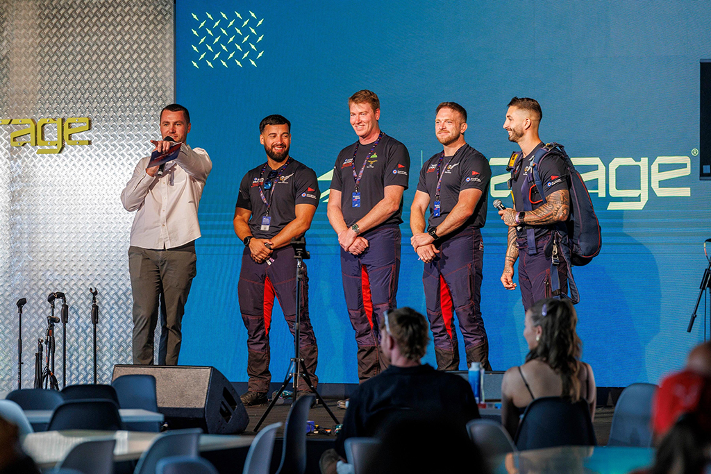 In the background, four Royal Air Force Falcons Parachute Display Team members, in conjunction with a compare, stand on a stage within a room, answering questions from a seated audience in the foreground. The Falcon on the righthand side is wearing a packed parachute for demonstration purposes.