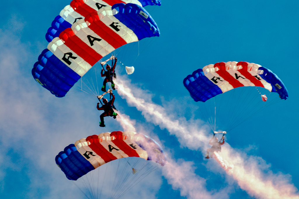 Four parachutists from the RAF Falcons parachute display team perform an aerial formation in a blue sky, trailing thick white smoke. Their rectangular parachutes feature a red, white, and blue striped pattern, with the letters "RAF" boldly printed across the central panels.