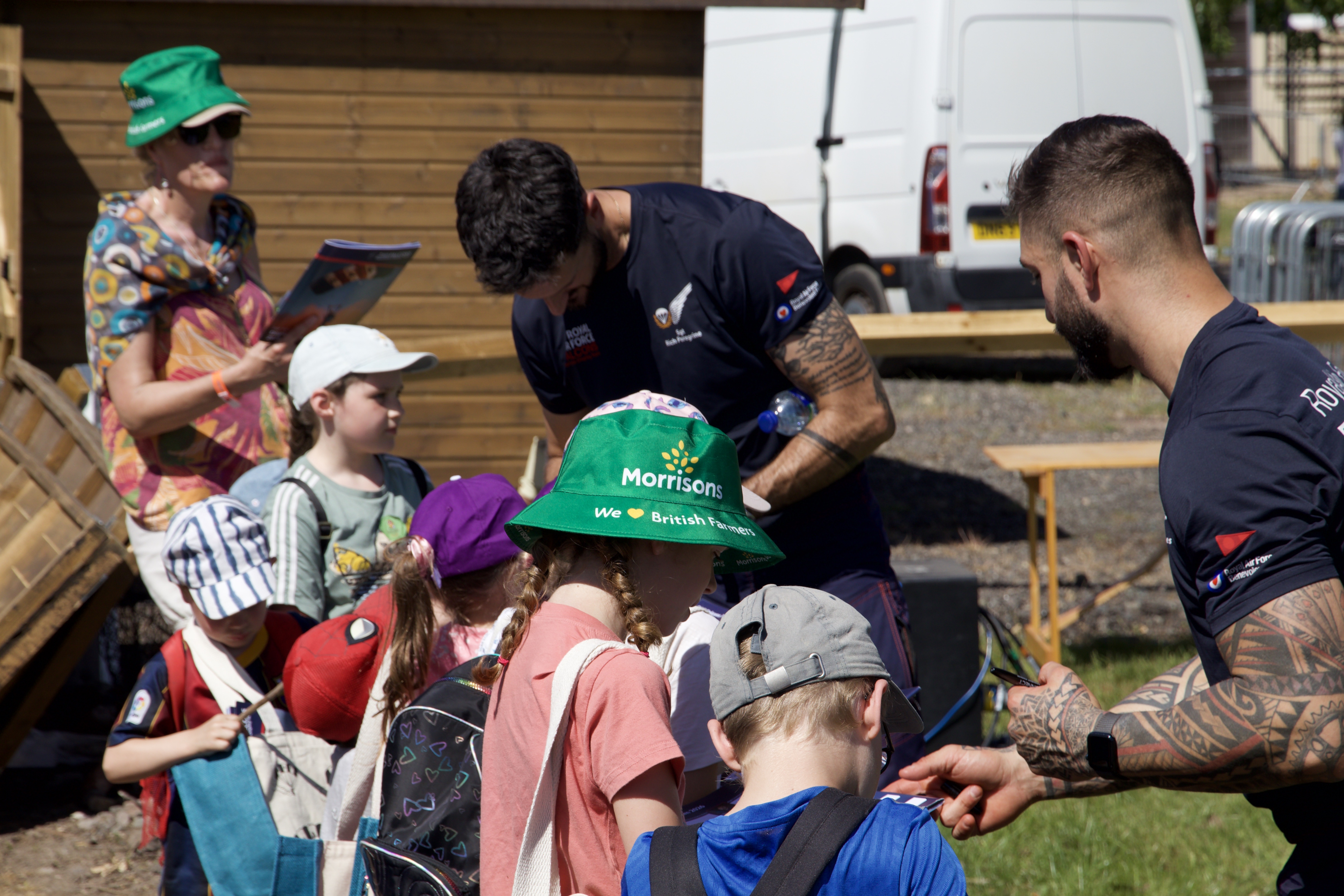 RAF Falcons team members interact with a group of children outdoors, post jump. One of the children wears a bright green bucket hat with "Morrisons We ❤ British Farmers" printed on it. In the background, a woman in a colorful shirt and green hat looks on, and a white van is partially visible.