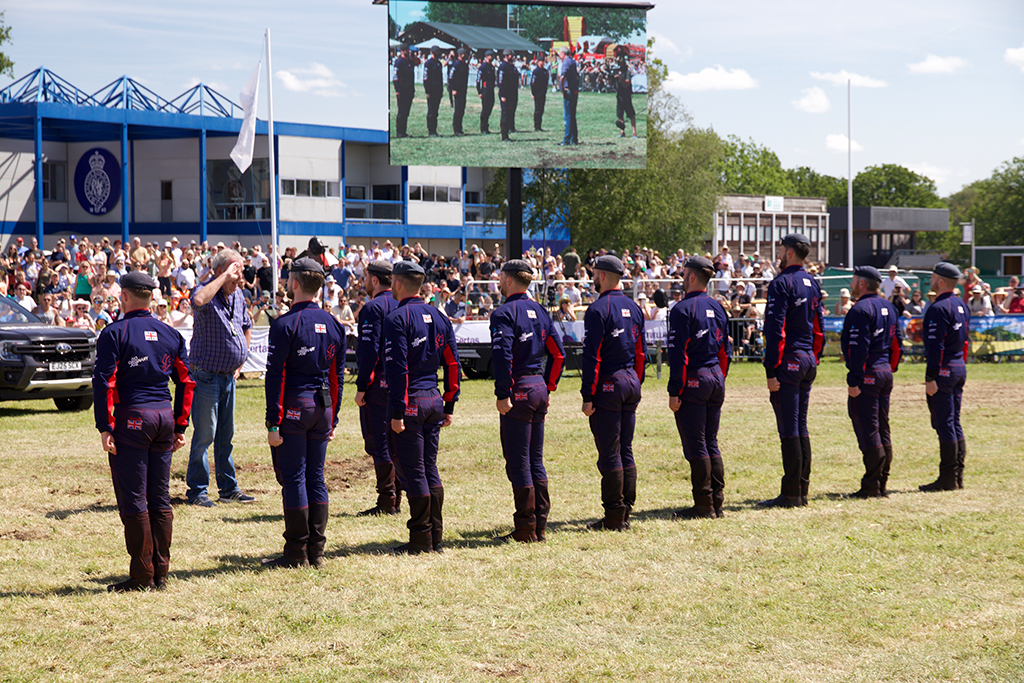 The RAF Falcons team in matching dark blue jumpsuits, stand at attention on a grassy field. Jeremy Clarkson stands before them, raising his hand in a salute. In the background, a large digital screen displays the scene to a seated crowd in front of a modern blue building under a clear sky.