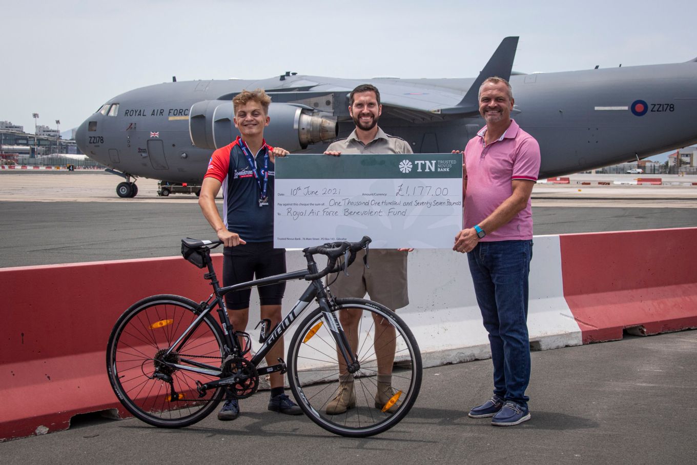 Oliver holds the cheque with his bicycle and a Globemaster in the background.