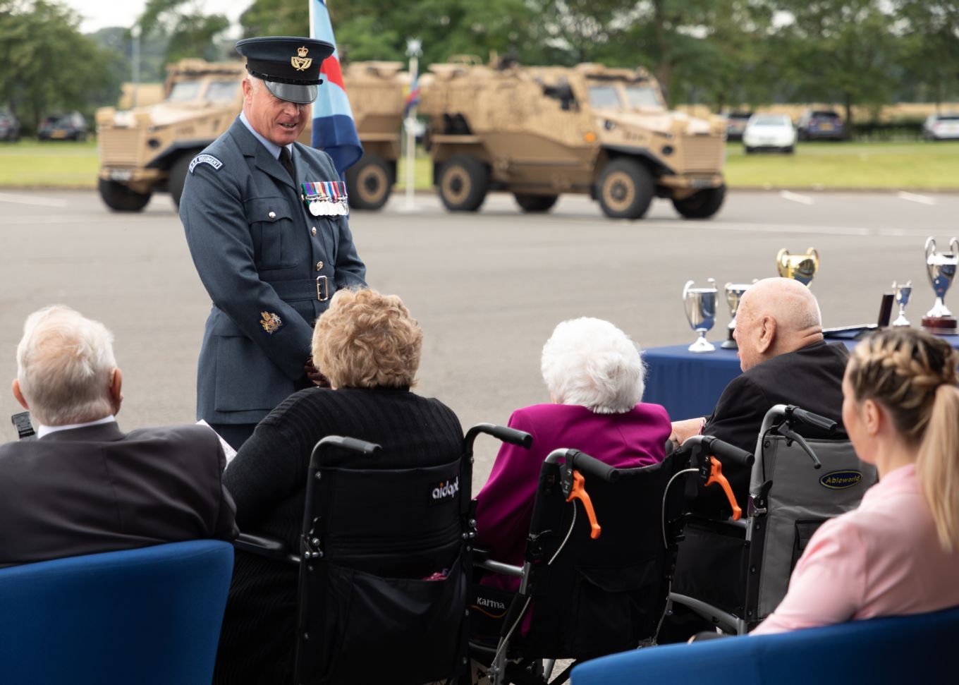 Audience with wheelchairs watch parade. Heavy vehicles in the background. 