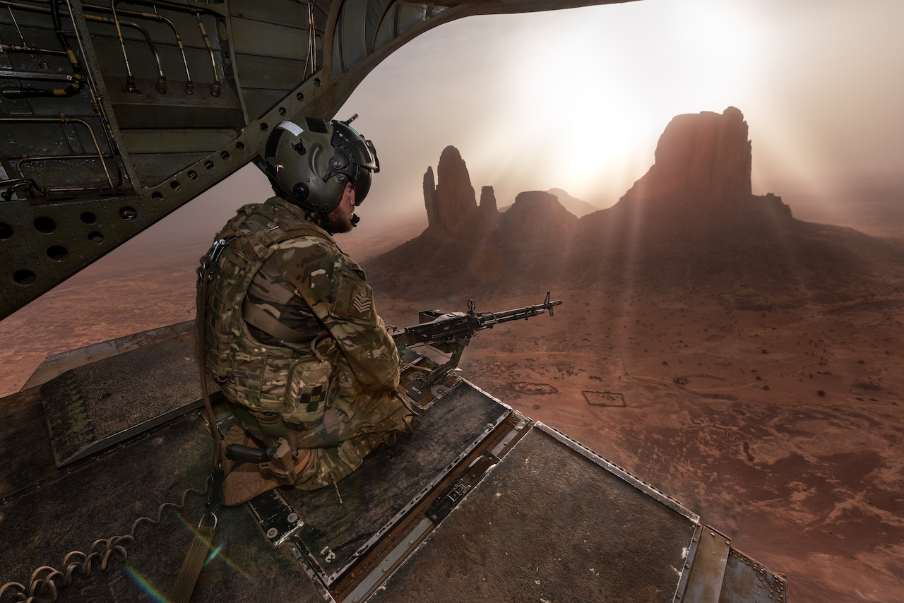 Rear door Gunner looks out from the Chinook. Sun rays break around landscape rock. 