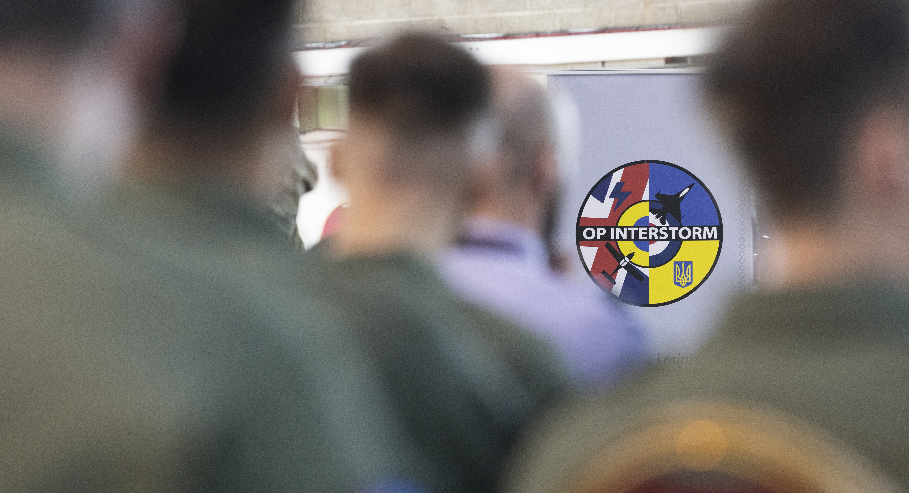 A blurred foreground of personnel frames a display board showing an “OP INTERSTORM” emblem.
