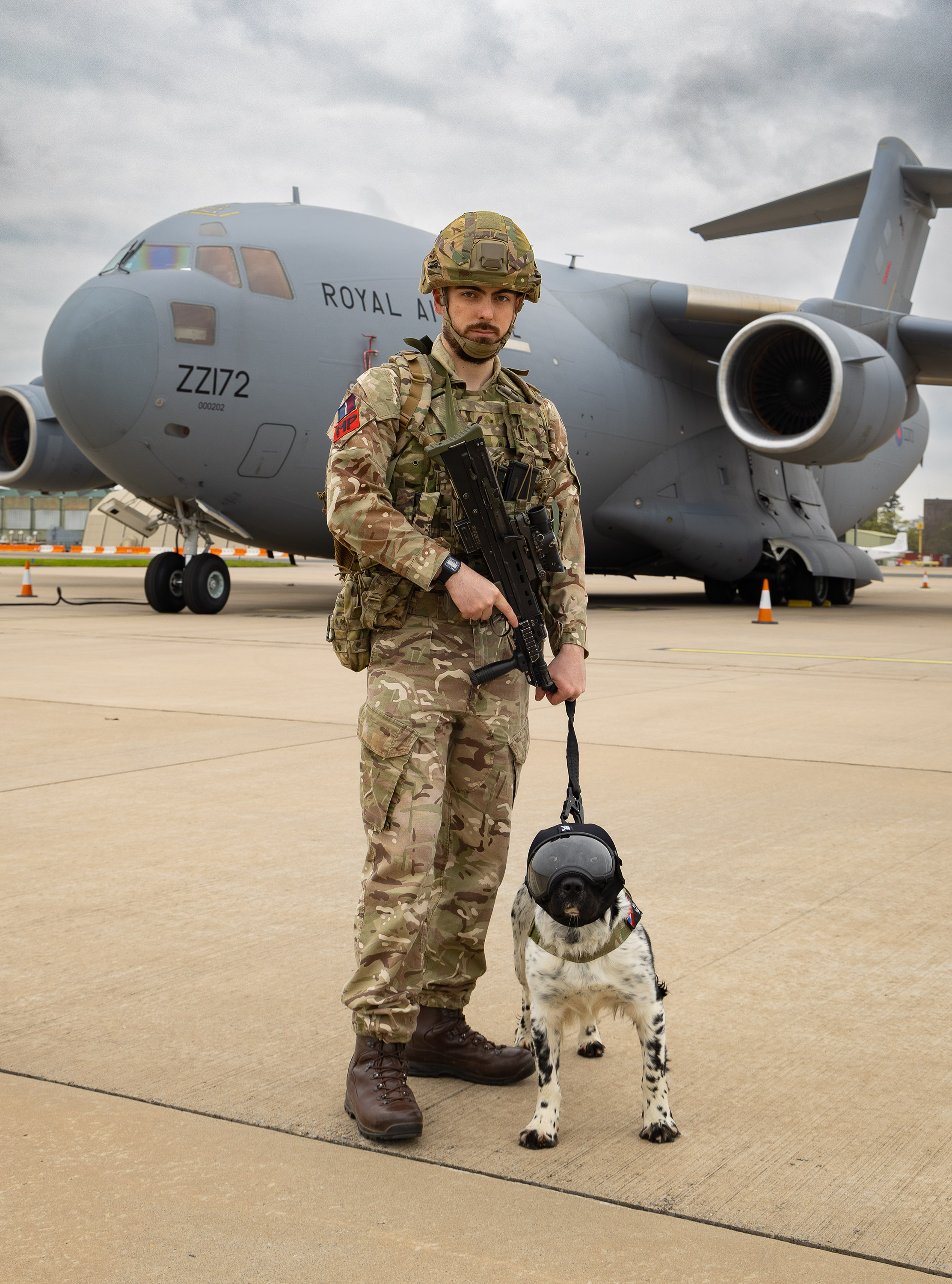 A member of the RAF stood next to a Military Working dog with an RAF A400M aircraft in the background.