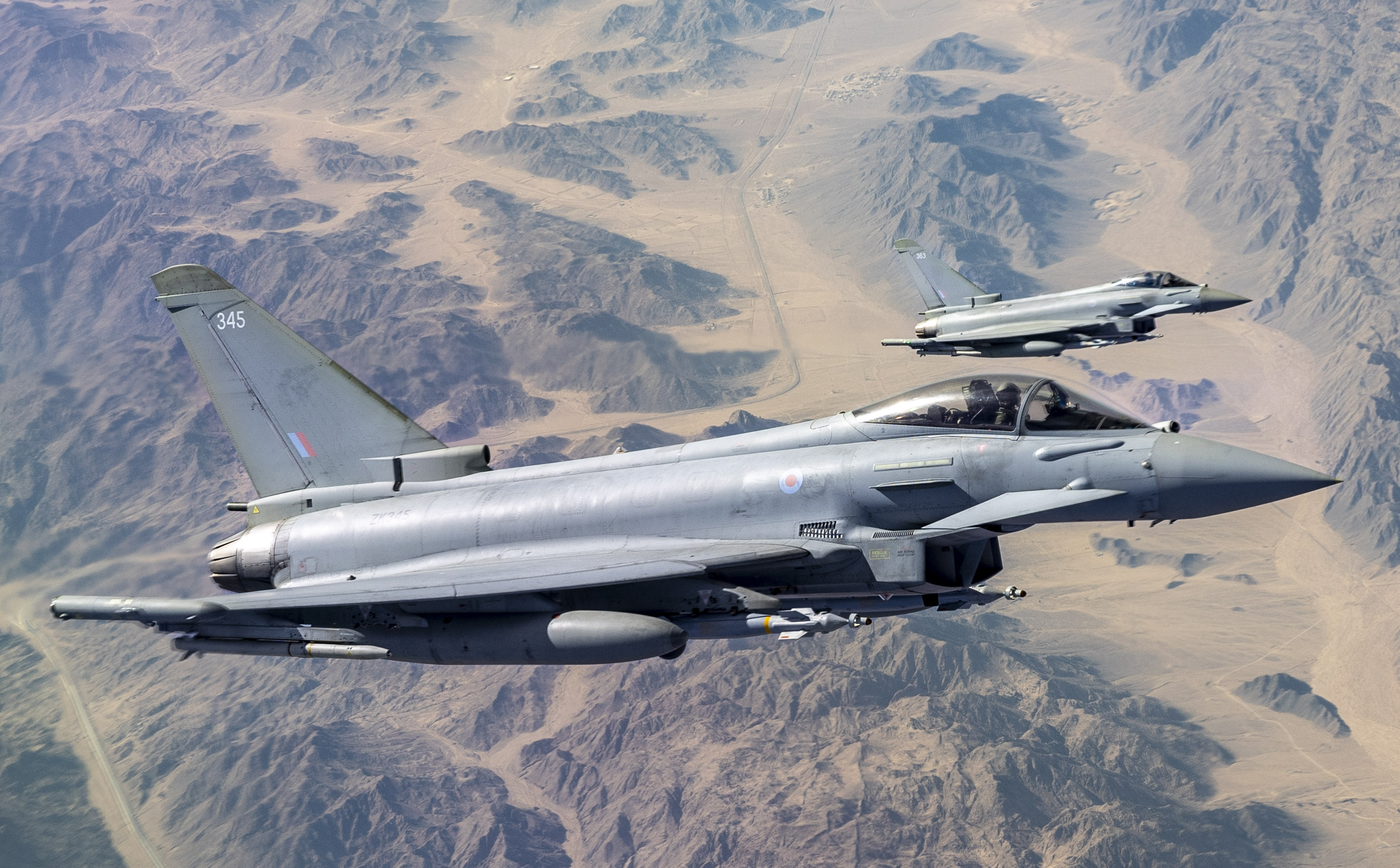 Two RAF Typhoons flying over a rugged rocky and sandy landscape.