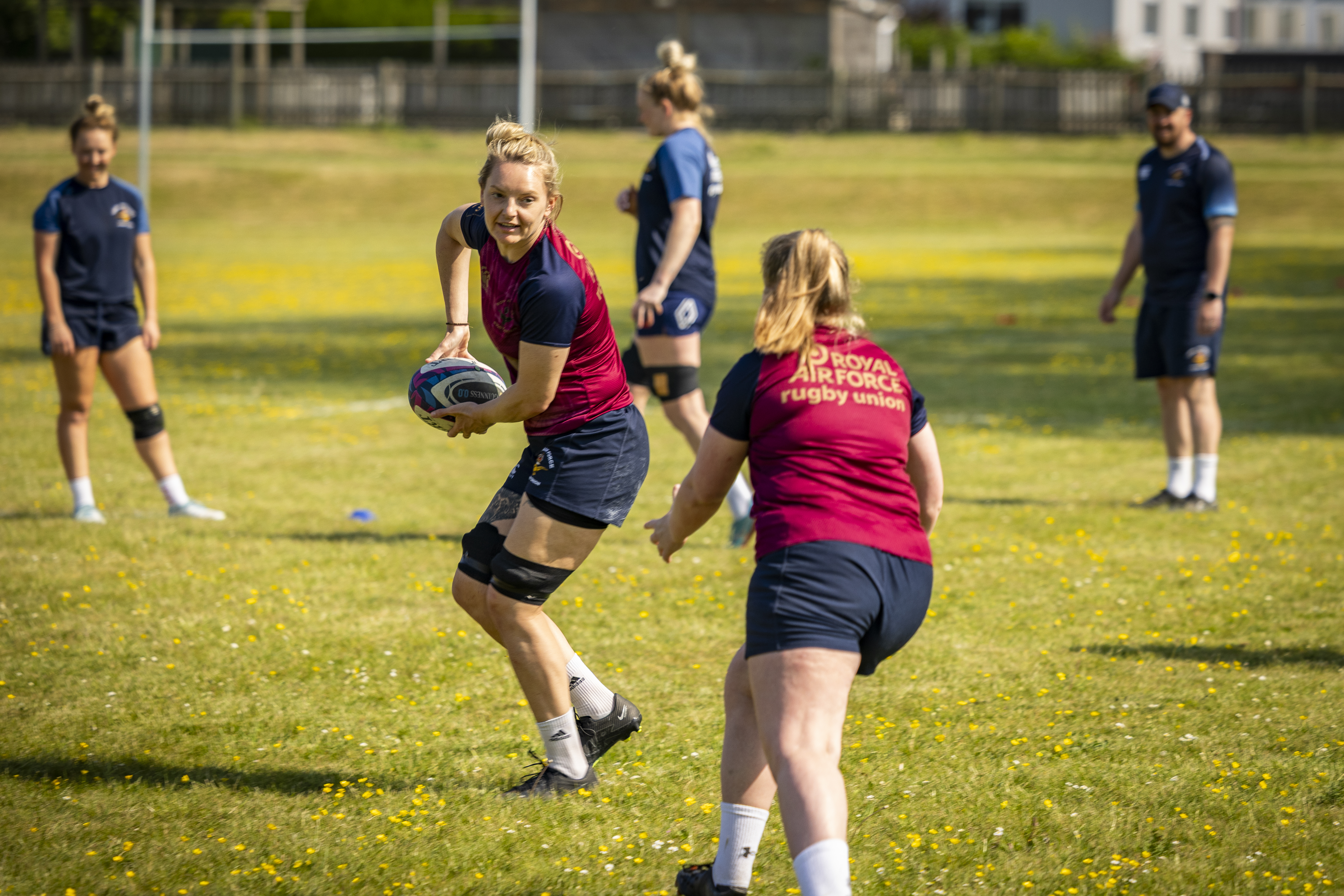 2 players passing a rugby ball between them, coaches stood in the background.