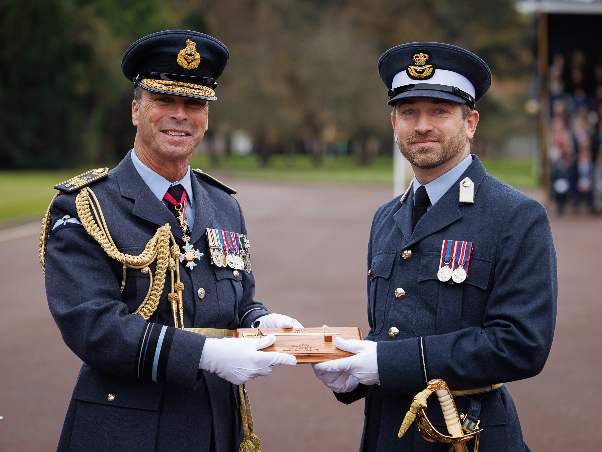Air Vice-Marshal and Officer Cadet both hold a miniature sword on wooden panel.