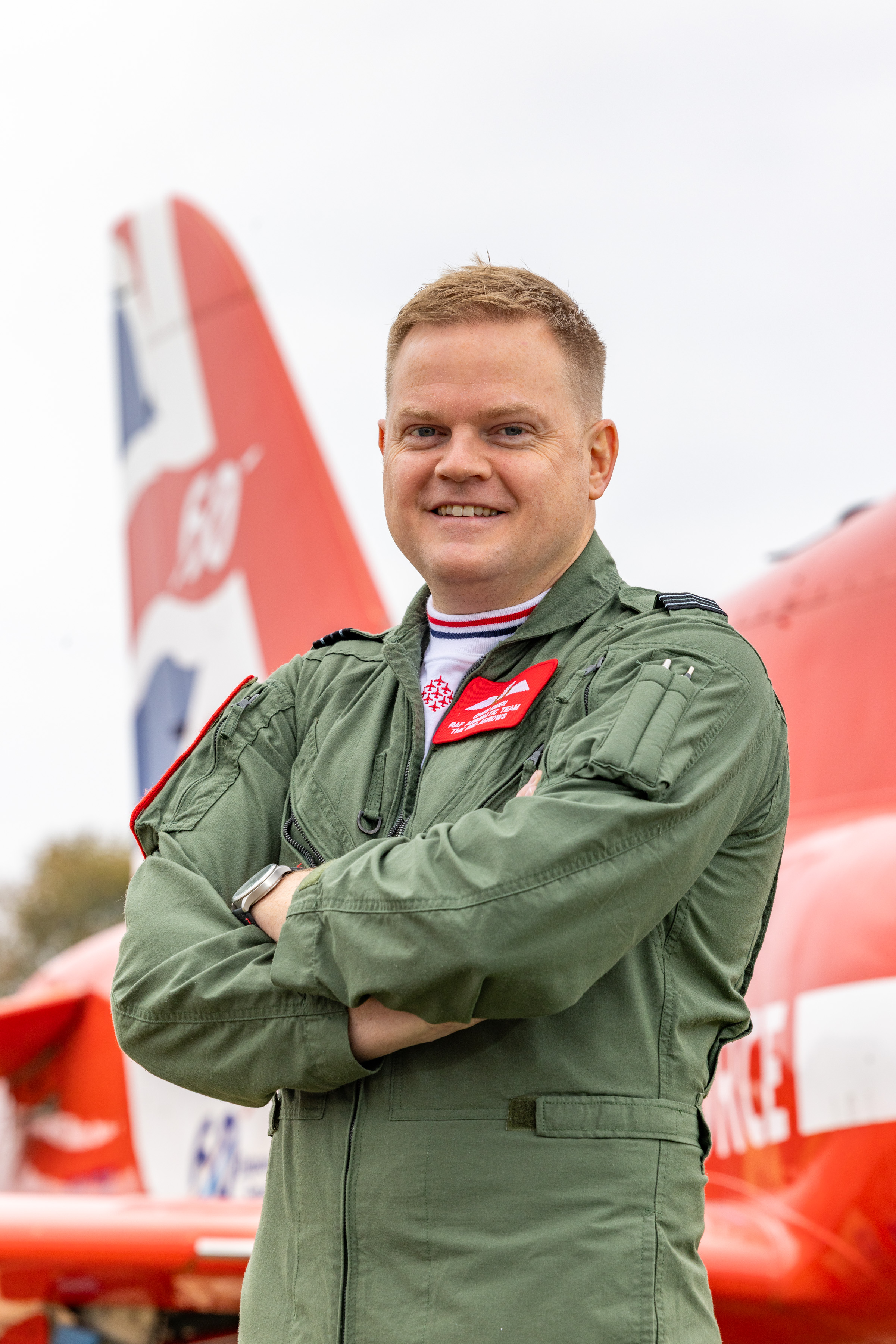 Joining for 2026 - this is Red Arrows pilot and Red 3 Flight Lieutenant Christopher Deen, wearing a green flying coverall, in front of a Red Arrows Hawk jet.
