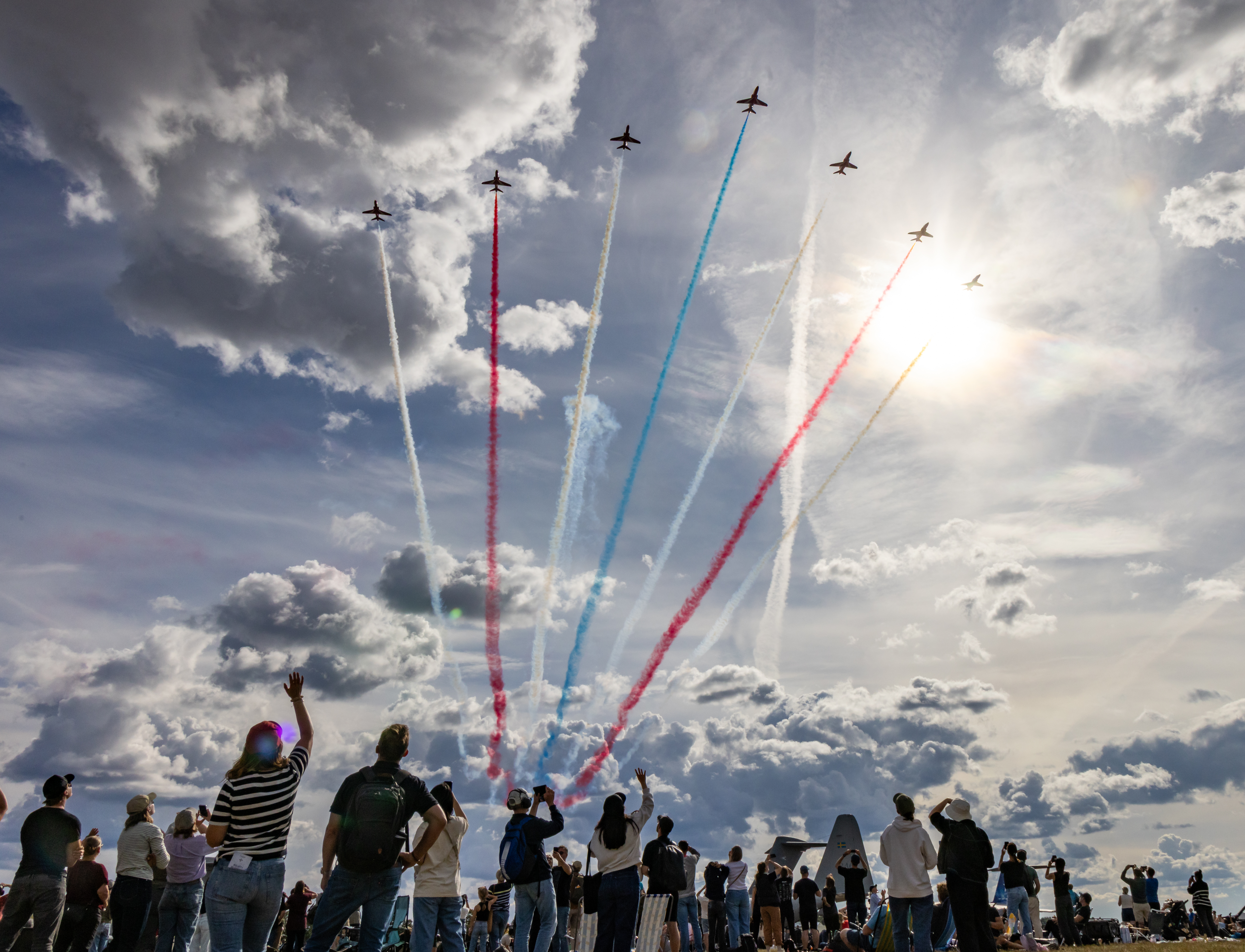 People at an airshow wave to the Red Arrows flying overhead in Cascade formation.