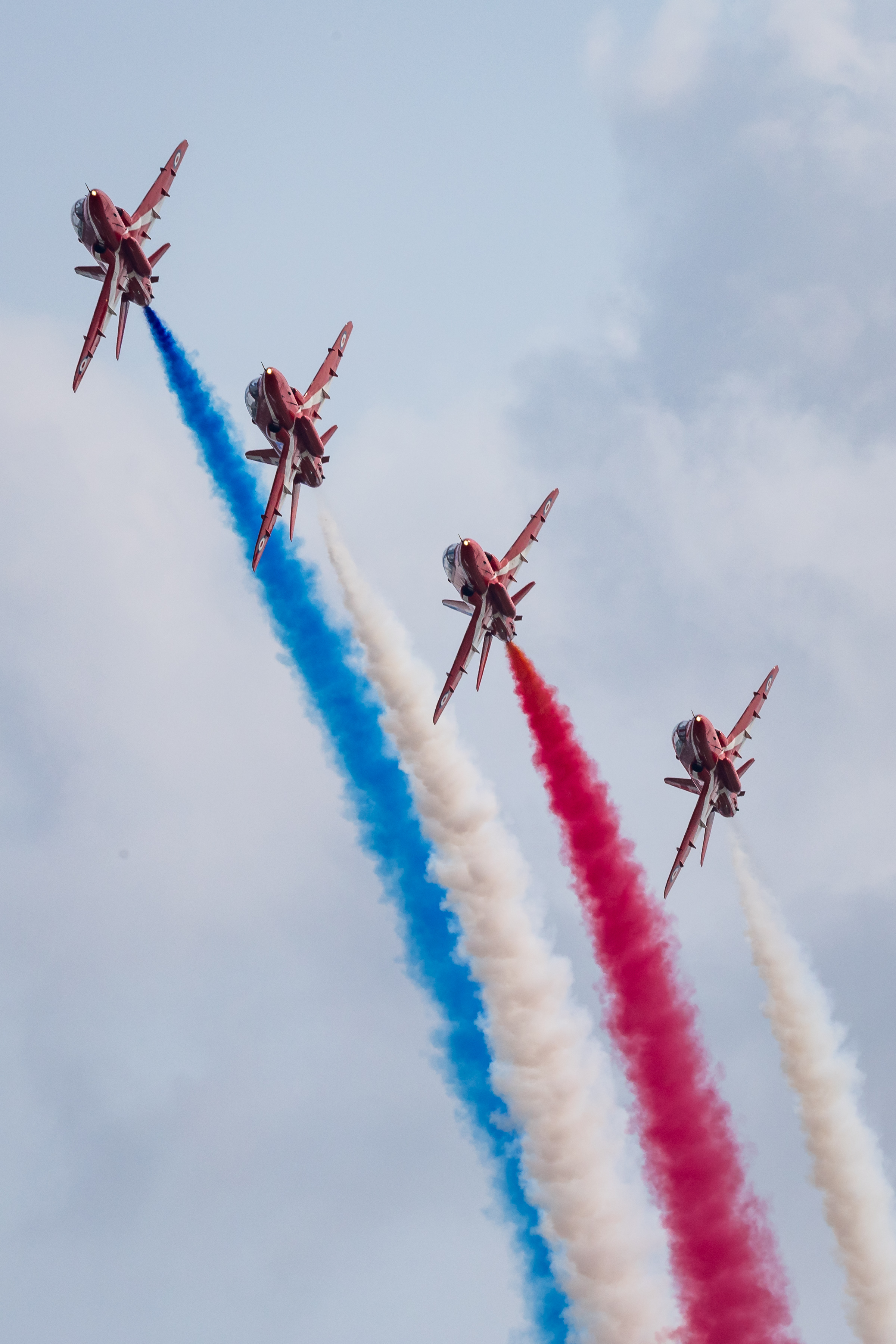 Four aircraft from the Red Arrows fly in close formation, stacked one above another, trailing red, white and blue smoke colours.