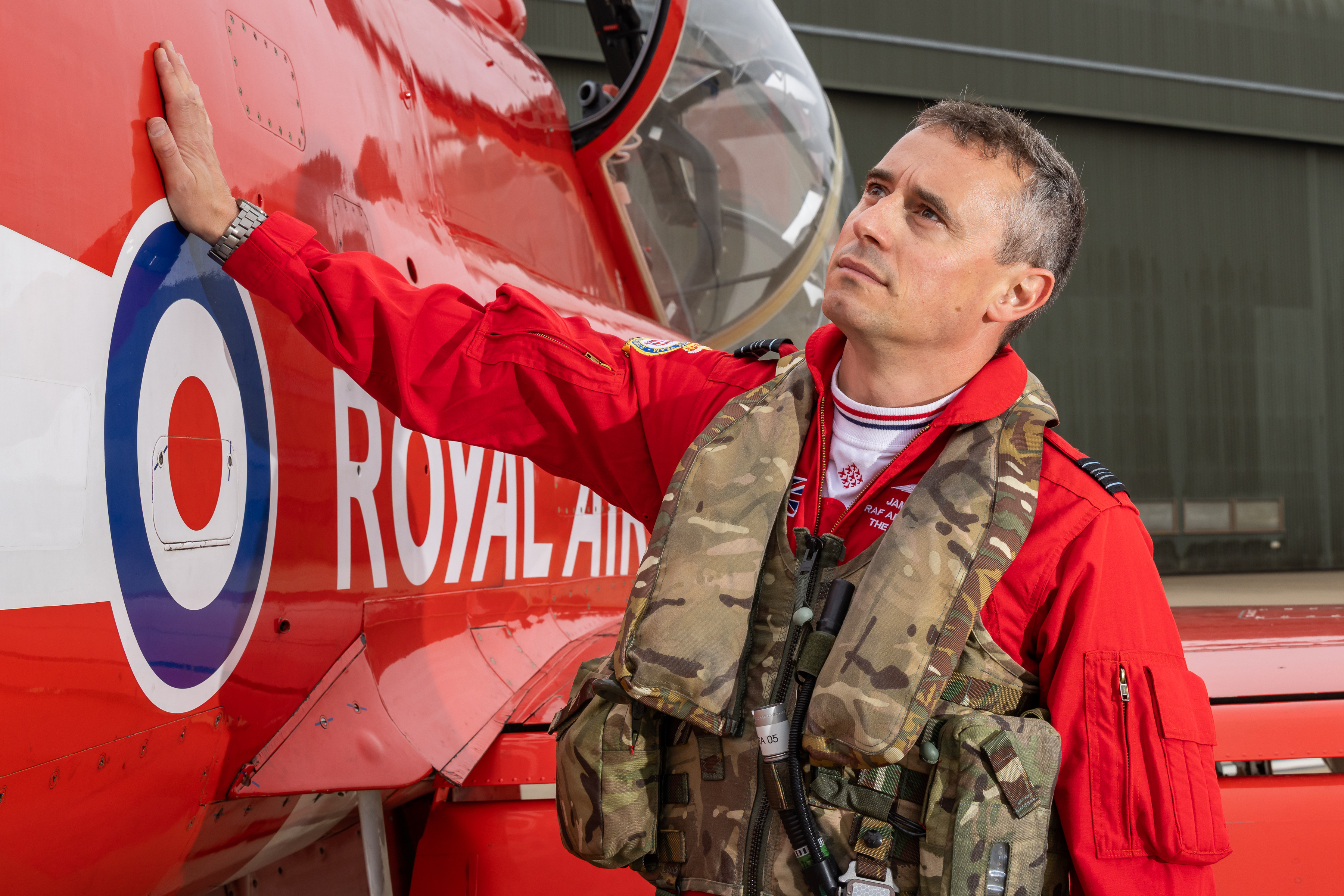 Squadron Leader James Turner, in a red flying suit, places his hand on a Red Arrows Hawk T1 jet and takes a closer look at the aircraft.