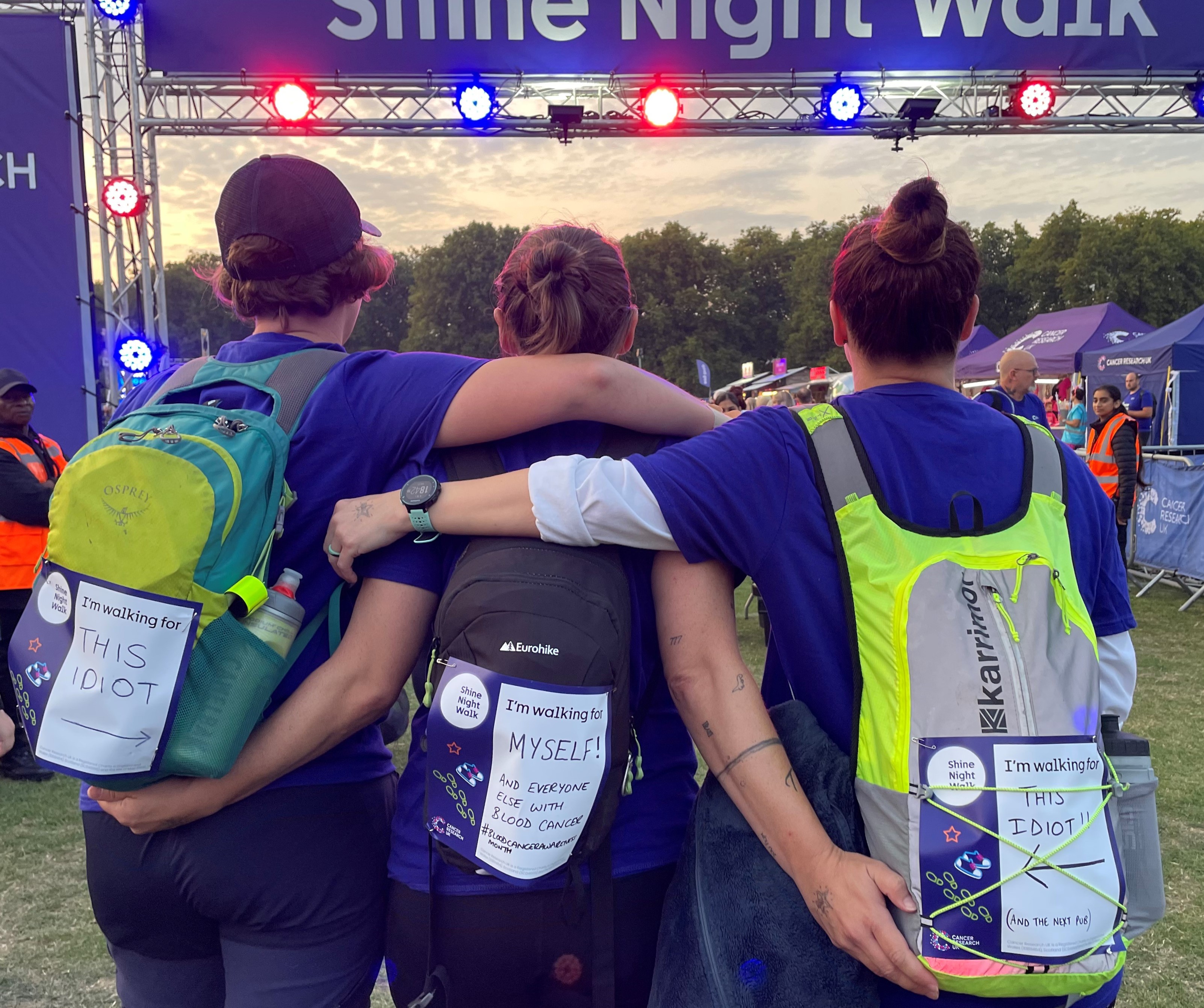 Three people hug eachother from behind with backpacks on.