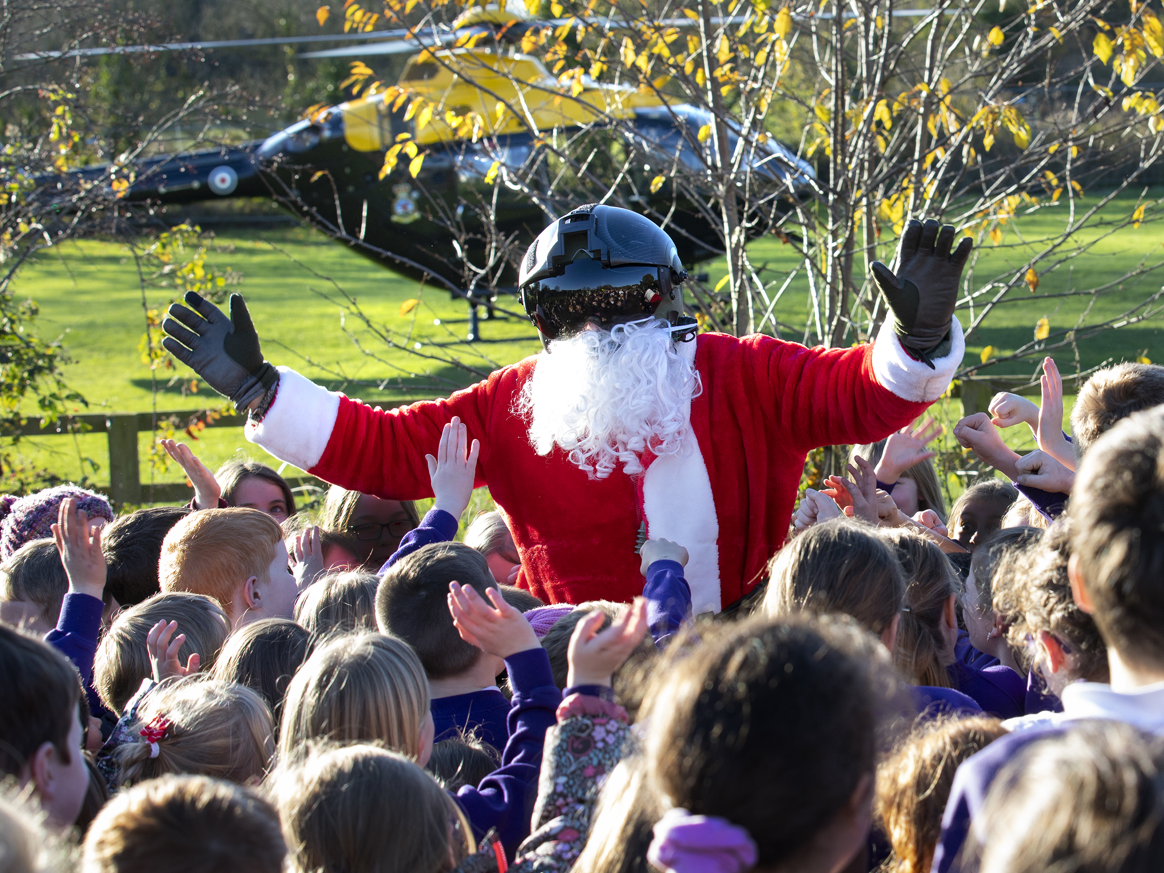 Santa Claus wearing a visor helmet and surrounded by schoolchildren, with a helicopter in the background.
