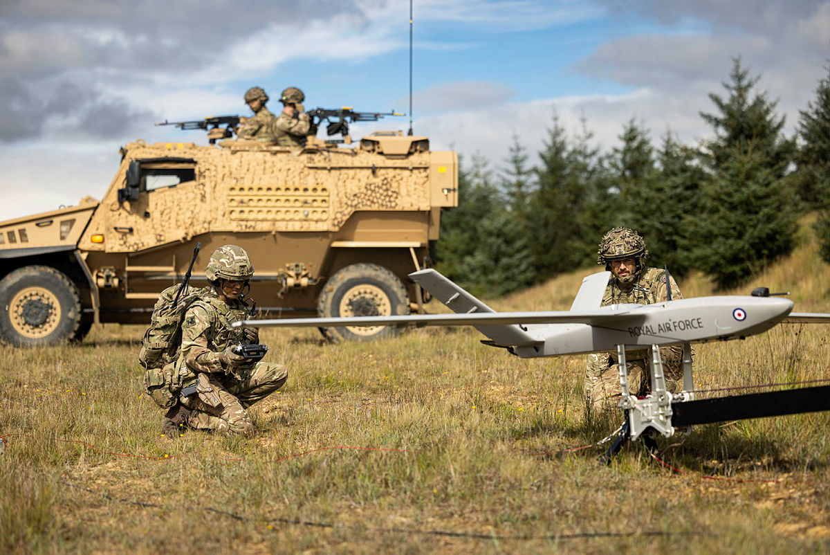 Two Royal Air Force personnel launching StormShroud while an armoured military vehicle is parked behind them in a field.