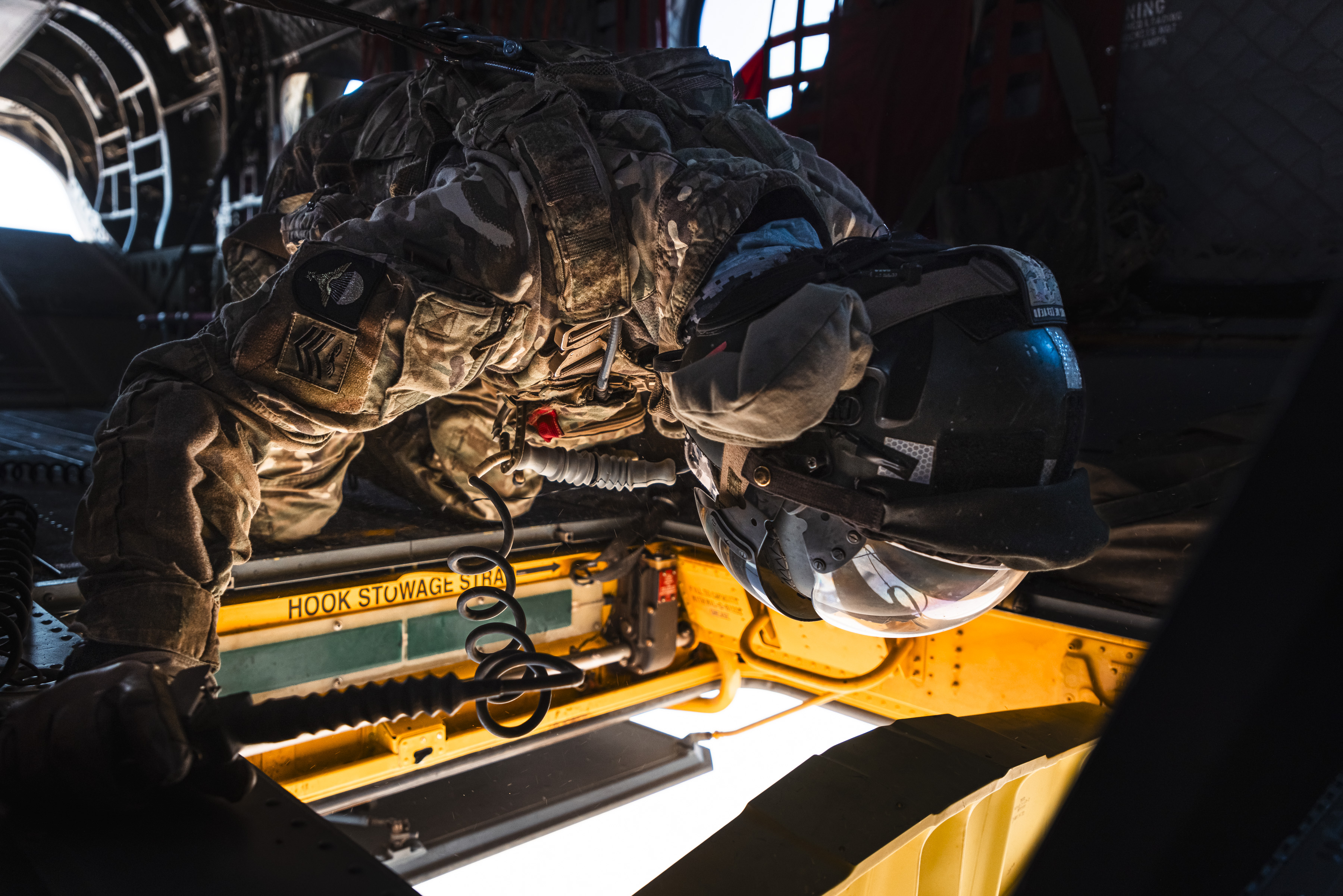 Member of Aircrew kneeling on floor of chinook, loooking down through the opened floor hatch.