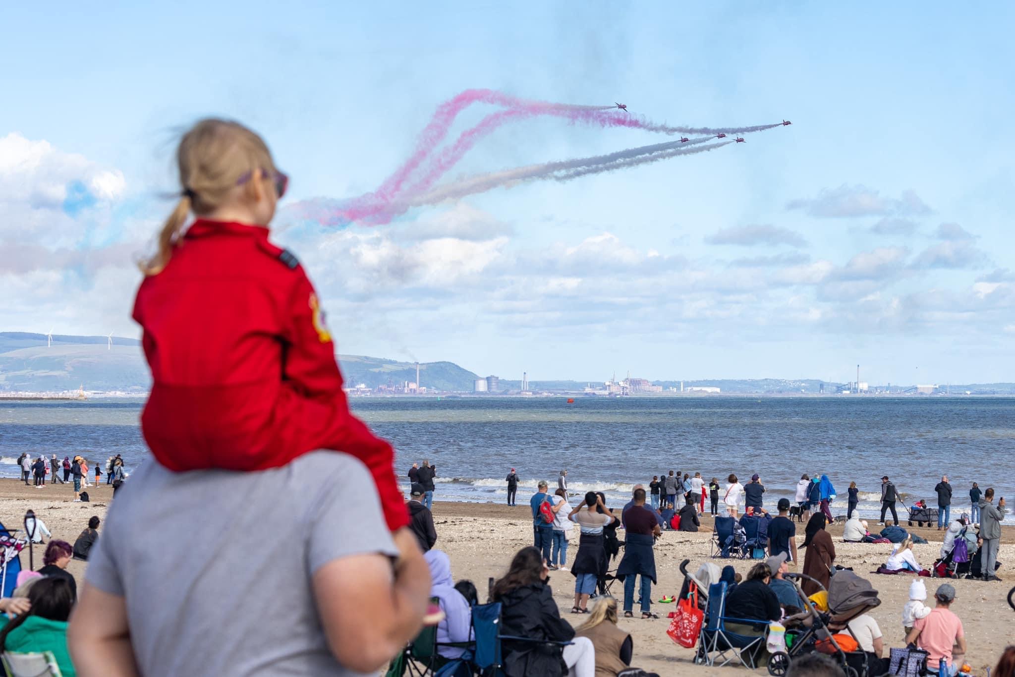 A child on an adult's shoulders watching the Red Arrows display in the sky.